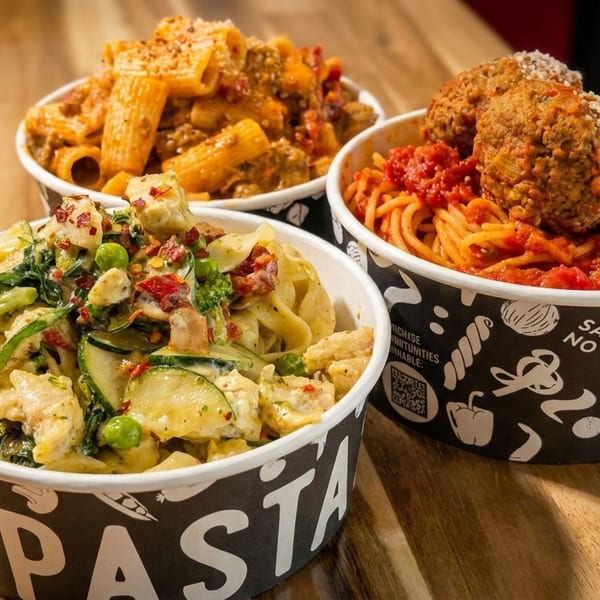 Three bowls of pasta on a wooden table: rigatoni with meat sauce, fettuccine with chicken and vegetables, and spaghetti with meatballs.