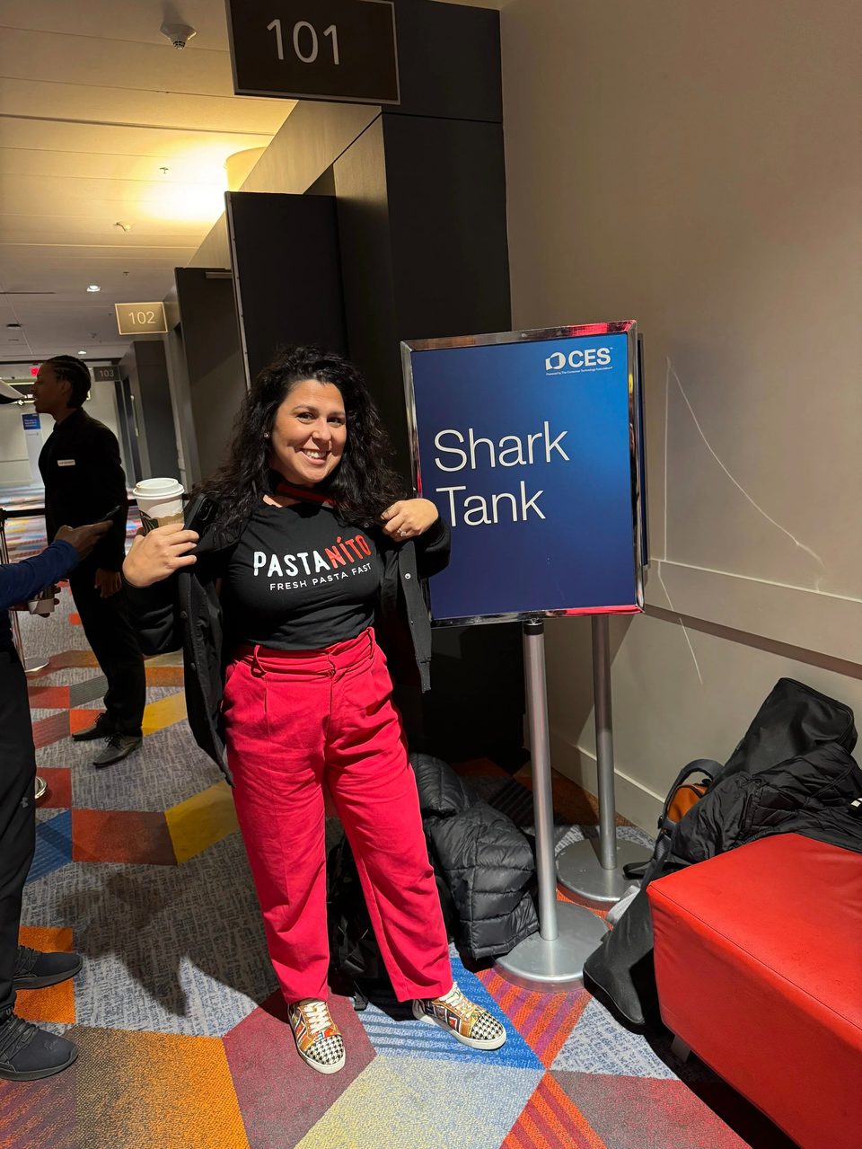 A woman in a "PASTANITO" shirt and red pants smiles next to a "Shark Tank" sign.