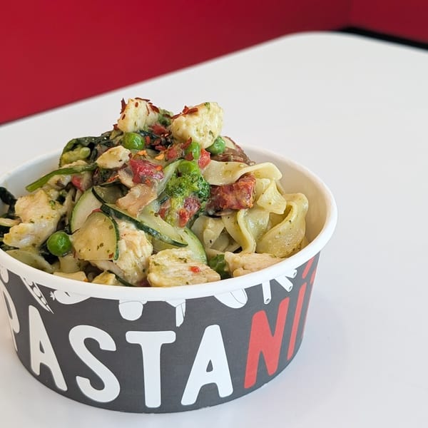 Pasta with chicken, zucchini, peas, and sun-dried tomatoes in a branded paper bowl.